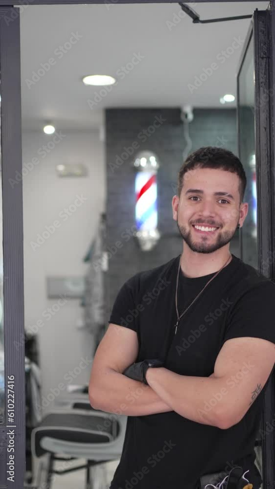 Vertical video portrait of a barber smiling at the camera leaning on the access door to his barbershop. Vertical video portrait of a barber smiling at the camera leaning on the access door to his barbershop.