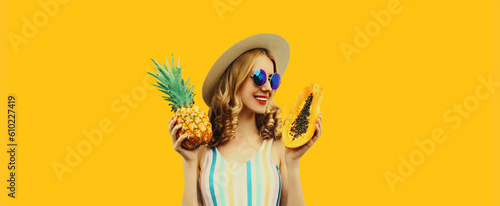 Summer portrait of happy cheerful smiling young woman with pineapple and papaya wearing straw hat, sunglasses on yellow background © rohappy