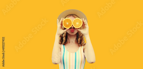 Summer portrait of cheerful funny young woman covering her eyes with fresh slices of orange fruits as binoculars looking for something wearing straw hat on yellow background