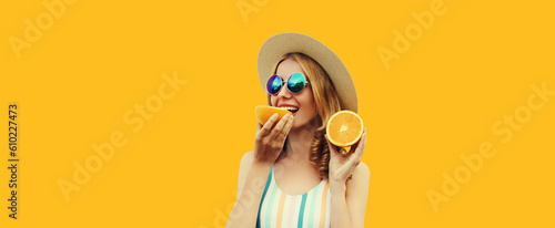 Summer portrait of happy young woman eating fresh juicy slices of orange fruits wearing straw hat, sunglasses on yellow background