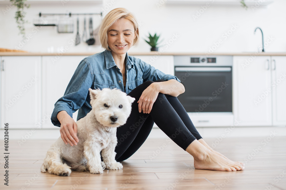 Portrait of cheerful young lady sitting next to well-behaved white dog on bare floor in kitchen interior. Affectionate female keeper giving canine friend overall attention on bright sunny day. Portrait of cheerful young lady sitting next to well-behaved white dog on bare floor in kitchen interior. Affectionate female keeper giving canine friend overall attention on bright sunny day.