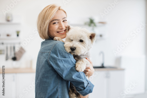 Fotografie Portrait of smiling blonde lady with West Highland White Terrier in arms posing on background of spacious kitchen