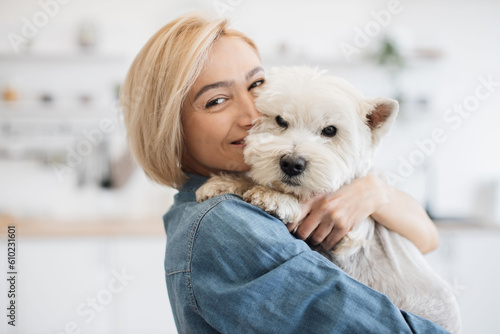 Obraz na plátně Portrait of smiling blonde lady with West Highland White Terrier in arms posing on background of spacious kitchen