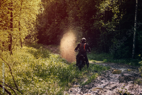 motorcycle racer on enduro sports motorcycle rides on a muddy road with puddles with splashes in forest in an off-road race on sunny day