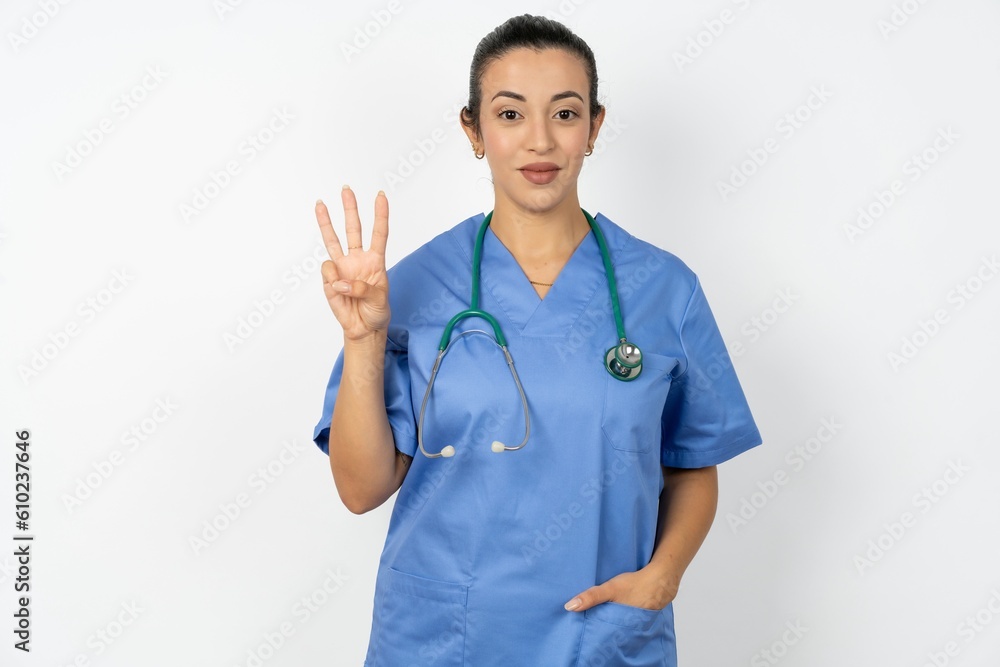 Young doctor woman wearing blue uniform over isolated background showing and pointing up with fingers number three while smiling confident and happy. Young doctor woman wearing blue uniform over isolated background showing and pointing up with fingers number three while smiling confident and happy.