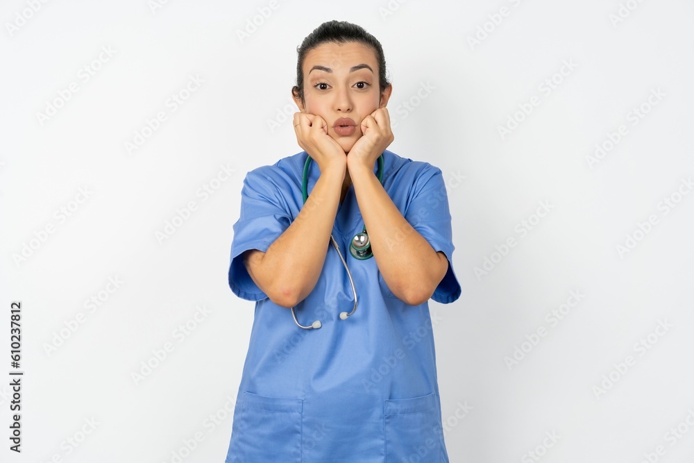 Young doctor woman wearing blue uniform over isolated background with surprised expression keeps hands under chin keeps lips folded makes funny grimace Young doctor woman wearing blue uniform over isolated background with surprised expression keeps hands under chin keeps lips folded makes funny grimace