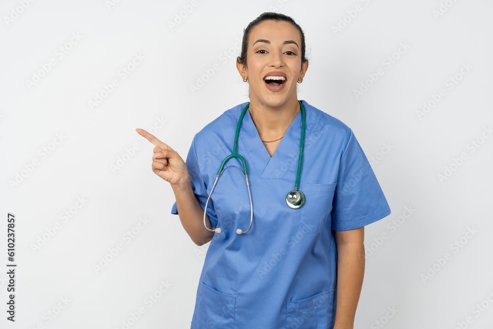 Young doctor woman wearing blue uniform over isolated background laughs happily points away on blank space demonstrates shopping discount offer, excited by good news or unexpected sale. Young doctor woman wearing blue uniform over isolated background laughs happily points away on blank space demonstrates shopping discount offer, excited by good news or unexpected sale.