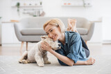 Pretty smiling woman in casual wear snuggling to cute pet's head while testing comfort of wooden floor with carpet on it. Joyful healthy lady appreciating affection and support of her canine friend.