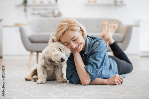 Kind female person and faithful small terrier snuggling to each other with heads while staying on wooden floor at home. Affectionate woman and cuddly Westie enjoying bonding interaction on sunny day.
