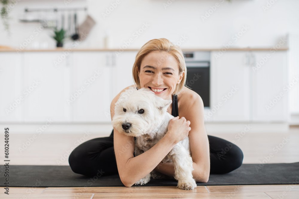 Affectionate adult female in yoga activewear embracing white terrier while sitting cross-legged on rubber mat indoors. Healthy lady awarding sweet pet with hug for good behaviour during home workout. Affectionate adult female in yoga activewear embracing white terrier while sitting cross-legged on rubber mat indoors. Healthy lady awarding sweet pet with hug for good behaviour during home workout.