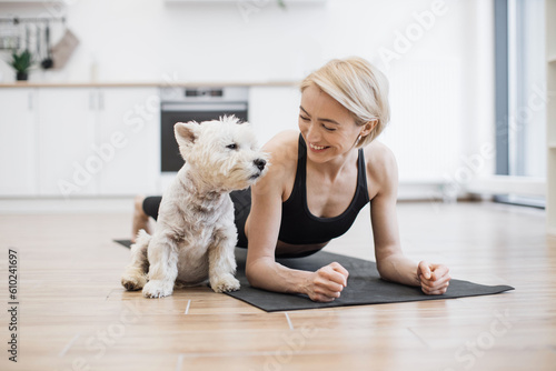 Φωτογραφία Healthy slim adult in sportswear holding arm balance yoga pose near white terrier sitting on black mat in kitchen