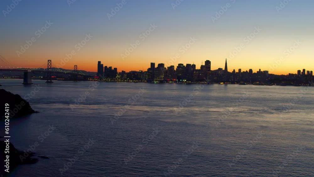 Aerial Shot Of Oakland Bay Bridge Near Skyscrapers In Downtown, Drone Descending Over Sea Near City Against Sky At Sunset - San Francisco, California Aerial Shot Of Oakland Bay Bridge Near Skyscrapers In Downtown, Drone Descending Over Sea Near City Against Sky At Sunset - San Francisco, California