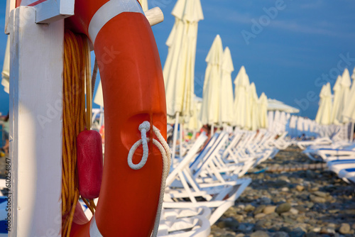 Lounge chairs with umbrellas lined up on clear rocky shore by sea against blue sky on beautiful beach. Lifebuoy and modern beach furniture located near the hotel poison with the sea for vacationers