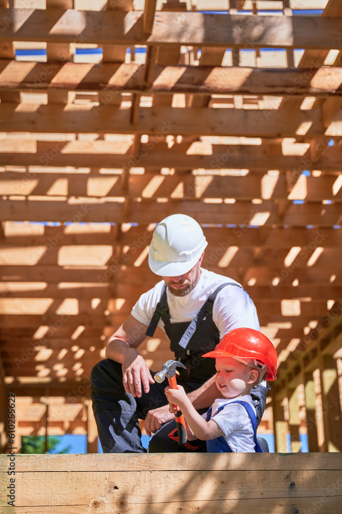 Father with toddler son constructing wooden frame house. Son attempting to hammer nail under the guidance of his father on construction site, wearing helmets and overalls. Carpentry and family concept Father with toddler son constructing wooden frame house. Son attempting to hammer nail under the guidance of his father on construction site, wearing helmets and overalls. Carpentry and family concept