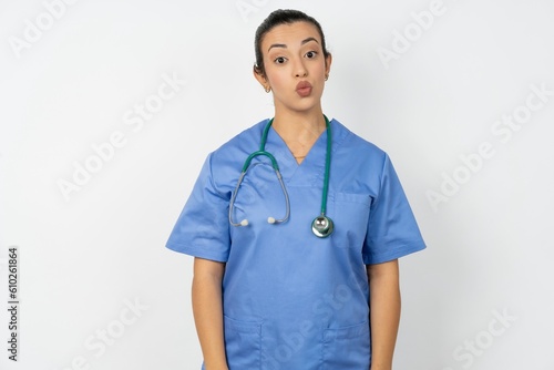 Shot of pleasant looking Beautiful doctor woman standing over white studio background, pouts lips, looks at camera, Human facial expressions