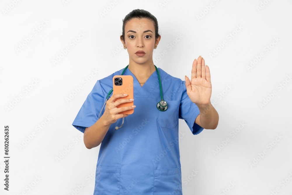 Beautiful doctor woman standing over white studio background using and texting with smartphone with open hand doing stop sign with serious and confident expression, defense gesture Beautiful doctor woman standing over white studio background using and texting with smartphone with open hand doing stop sign with serious and confident expression, defense gesture
