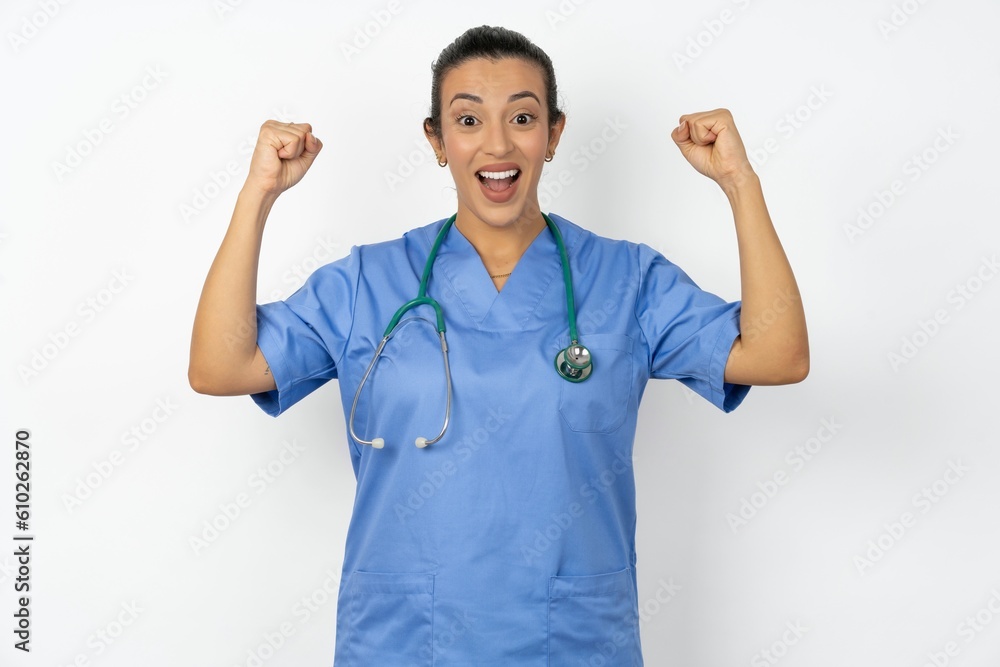 Shocked ecstatic Beautiful doctor woman standing over white studio background win luck lottery raise hands up shout yea