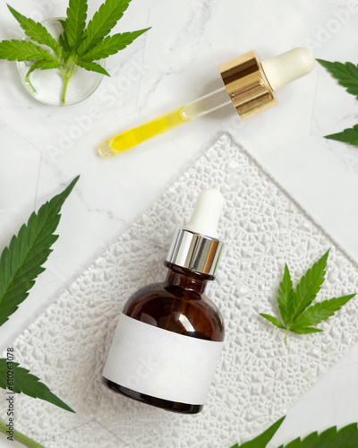 Dropper bottle with blank label near green cannabis leaves on white table. Cosmetic Mockup