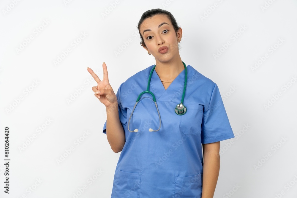 Beautiful doctor woman standing over white studio background makes peace gesture keeps lips folded shows v sign. Body language concept Beautiful doctor woman standing over white studio background makes peace gesture keeps lips folded shows v sign. Body language concept