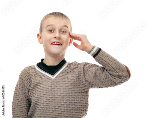 Portrait of blonde year old boy saluting with grimace on his face and smiling to camera, isolated on white background. Attractive child in brown sweater holding hand on his forehead posing in studio.
