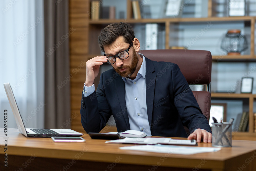 Upset businessman behind paper work inside modern office, mature man with beard reading financial reports and account documents unhappy with results and disappointed with achievements. Upset businessman behind paper work inside modern office, mature man with beard reading financial reports and account documents unhappy with results and disappointed with achievements.
