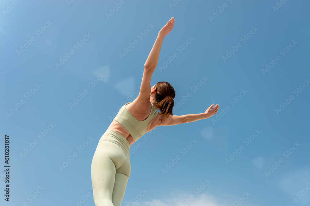 Fototapeta premium Rear view of a sporty woman with her arms raised in success, against a blue sky.