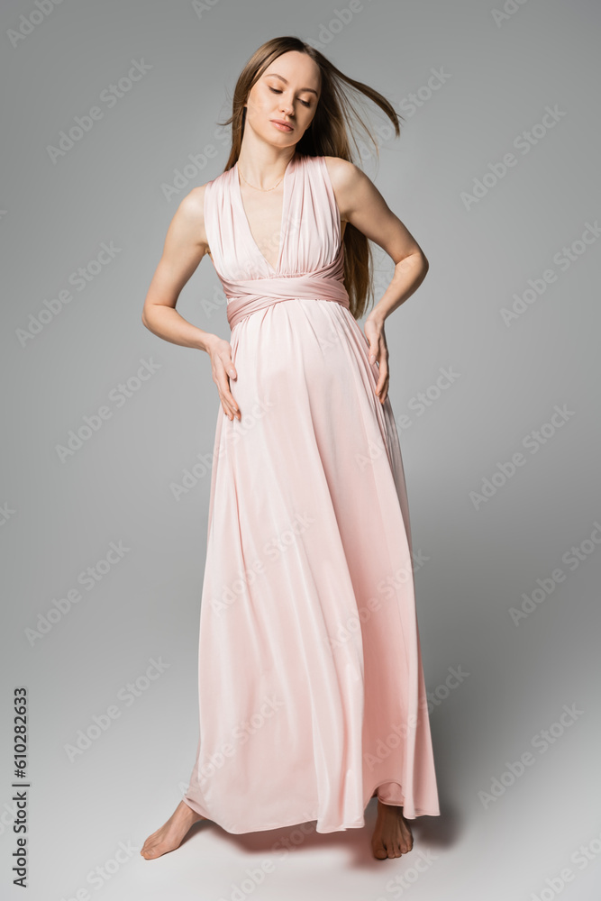 Full length of barefoot long haired and pregnant woman in pink dress touching belly while standing on grey background, elegant and stylish pregnancy attire, sensuality, mother-to-be Full length of barefoot long haired and pregnant woman in pink dress touching belly while standing on grey background, elegant and stylish pregnancy attire, sensuality, mother-to-be