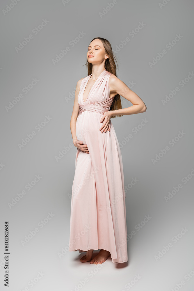 Full length of relaxed and fair haired pregnant woman in pink dress touching belly while standing on grey background, elegant and stylish pregnancy attire, sensuality, mother-to-be Full length of relaxed and fair haired pregnant woman in pink dress touching belly while standing on grey background, elegant and stylish pregnancy attire, sensuality, mother-to-be