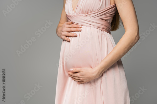 Cropped view of fashionable and long haired woman in pink dress touching belly while standing isolated on grey, elegant and stylish pregnancy attire, sensuality, mother-to-be