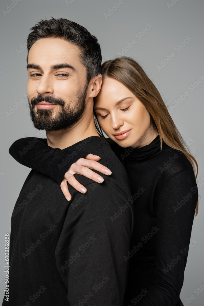 Portrait of relaxed fair haired woman in black clothes hugging bearded husband while standing together isolated on grey, husband and wife relationship concept Portrait of relaxed fair haired woman in black clothes hugging bearded husband while standing together isolated on grey, husband and wife relationship concept