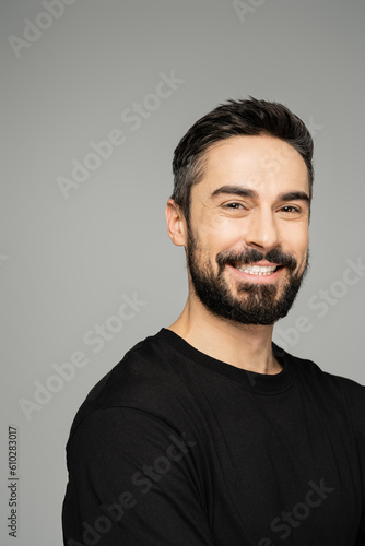Portrait of confident and cheerful bearded man in casual black t-shirt looking at camera and standing isolated on grey, masculine beauty concept