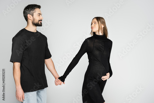 Smiling bearded man in black t-shirt holding hand of fair haired and pregnant wife in stylish dress on grey background, new beginnings and anticipation concept, expecting parents