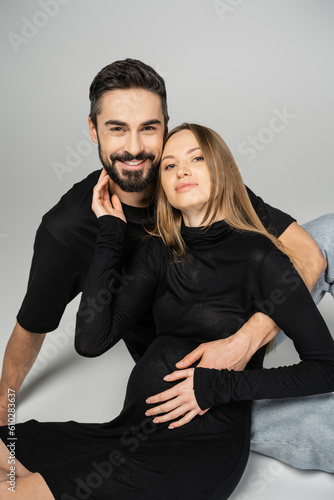 Cheerful bearded man in black t-shirt hugging trendy pregnant wife in dress and looking at camera while sitting on grey background, new beginnings and parenting concept © LIGHTFIELD STUDIOS