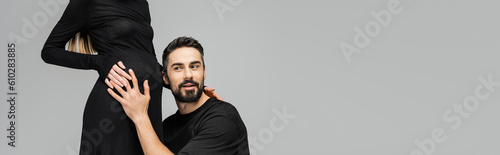 Excited and bearded husband in t-shirt listening and touching belly of pregnant and stylish wife in black dress isolated on grey with copy space, growing new life concept, banner