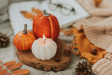 Cozy autumn decor - burning candle shape of pumpkin and orange decor pumpkins on wooden board on bed with open book, warm sweater, autumn leaves