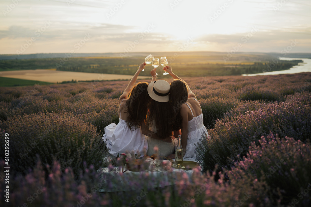 Back view of company of young female friends with hands up having fun, raising glasses with wine and enjoy beautiful sunset at summer picnic in lavender field.