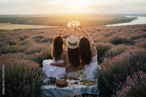 Young female friends having fun, raising glasses with wine and enjoy beautiful sunset at summer picnic in lavender field.	
