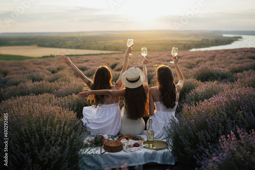 Girlfriends having picnic in the lavender field at sunset. Group of young women sitting on lavender field on summer day. Girlfriends drinking wine on outdoor party.	
