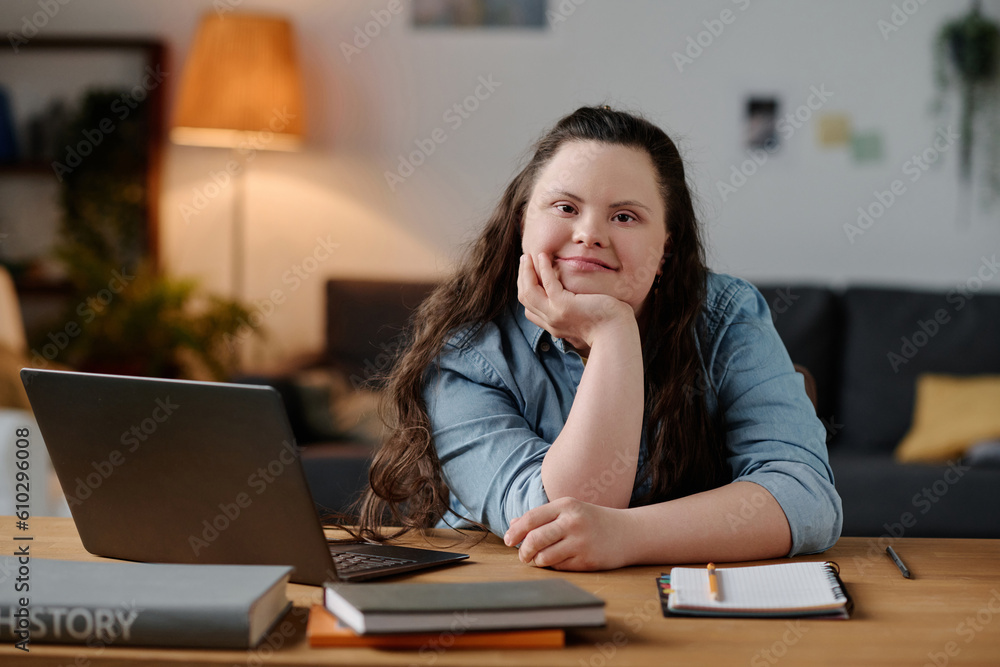Portrait of girl with down syndrome looking at camera while sitting at desk with laptop in the living room Portrait of girl with down syndrome looking at camera while sitting at desk with laptop in the living room