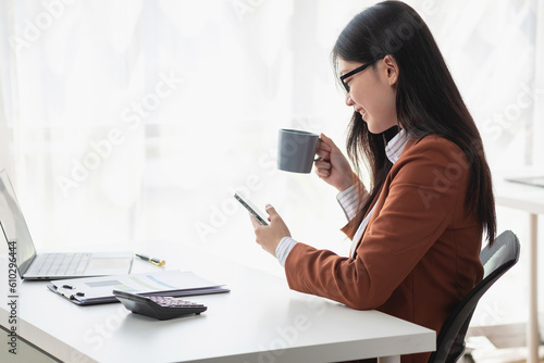 Young businesswoman with cheerful face sitting drinking coffee and using laptop in office Check email on the computer, sitting at a desk. Interior of the modern office.