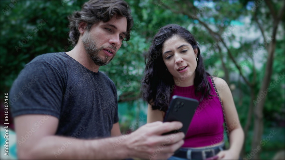 Young man showing online content to woman on cellphone device, both standing outside at city park. Two people looking at smartphone screen comenting on the media they are watching Young man showing online content to woman on cellphone device, both standing outside at city park. Two people looking at smartphone screen comenting on the media they are watching