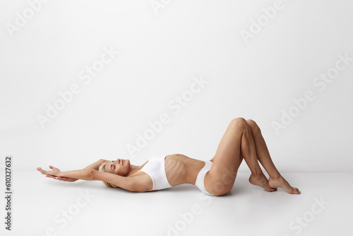 Self-love and care. Young woman with blonde short hair, skin body lying on floor, posing in underwear against grey studio background