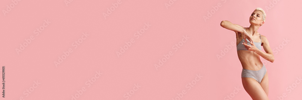 Naklejka premium Portrait of tender, young blonde woman with slim body posing i cotton grey underwear against pink studio background