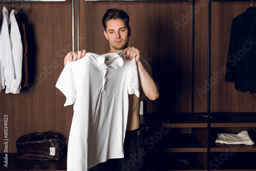 Handsome shirtless slim young man trying on white polo shirt in dressing room