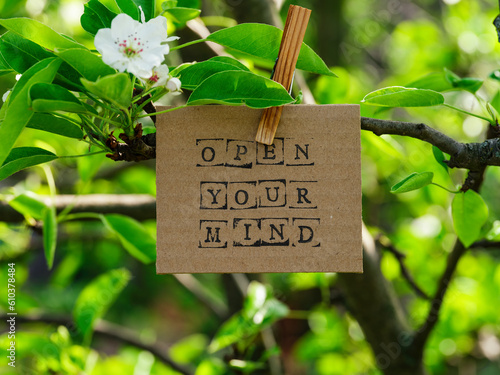 Piece of cardboard with words Open Your Mind on it hanging on a pear tree branch with blossoms and leaves using a wooden clothespin. © rosinka79