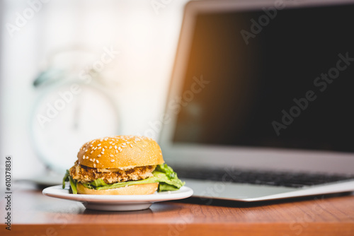 A delicious hamburger on the table with a blurred computer laptop and an alarm clock for eating fast food at work concept
