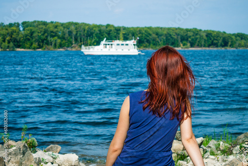 A traveler girl with red hair looks at a ship in the river from behind