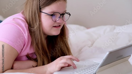 Focused overweight teenager looking at screen of tablet using touchpad on keyboard girl in black-rimmed glasses doing online exercises on website lying on bed at home photo