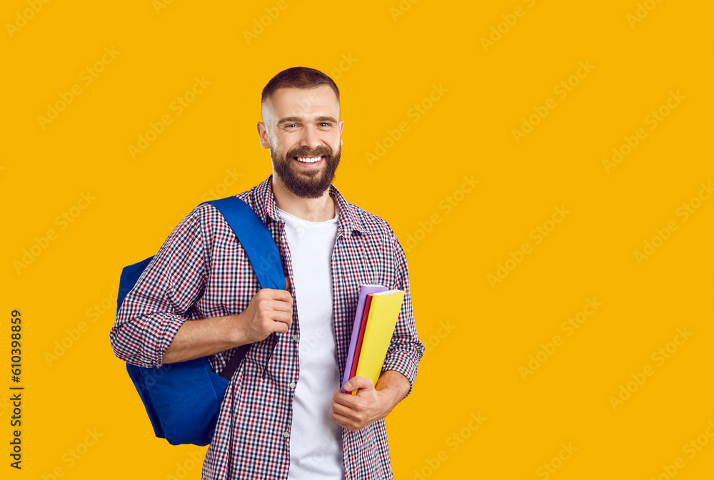Portrait of happy male college or university student with notebooks and backpack. Cheerful handsome young man in casual shirt standing on yellow orange copy space background, holding books and smiling Portrait of happy male college or university student with notebooks and backpack. Cheerful handsome young man in casual shirt standing on yellow orange copy space background, holding books and smiling