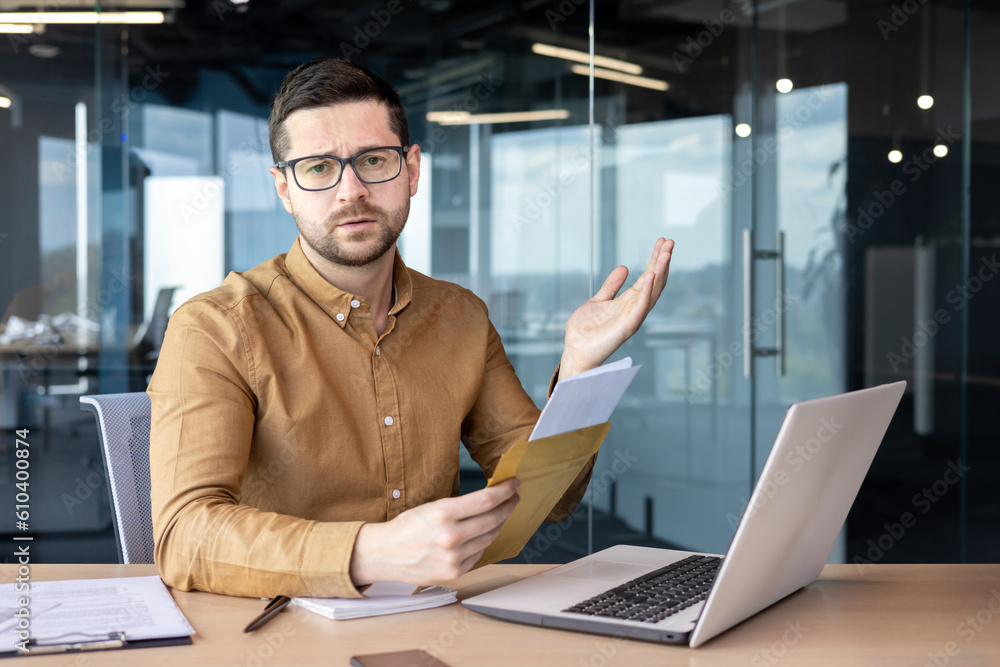 Disappointed young man businessman received bad news, information through letter. He sits at the desk in the office and holds an envelope, spreads his hands in frustration towards the camera Disappointed young man businessman received bad news, information through letter. He sits at the desk in the office and holds an envelope, spreads his hands in frustration towards the camera
